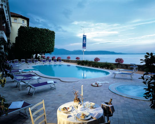 Grand Hotel Gardone view of the pool with blue sun beds set up surrounding it and a circular plunge poor on the side with the lake in the background