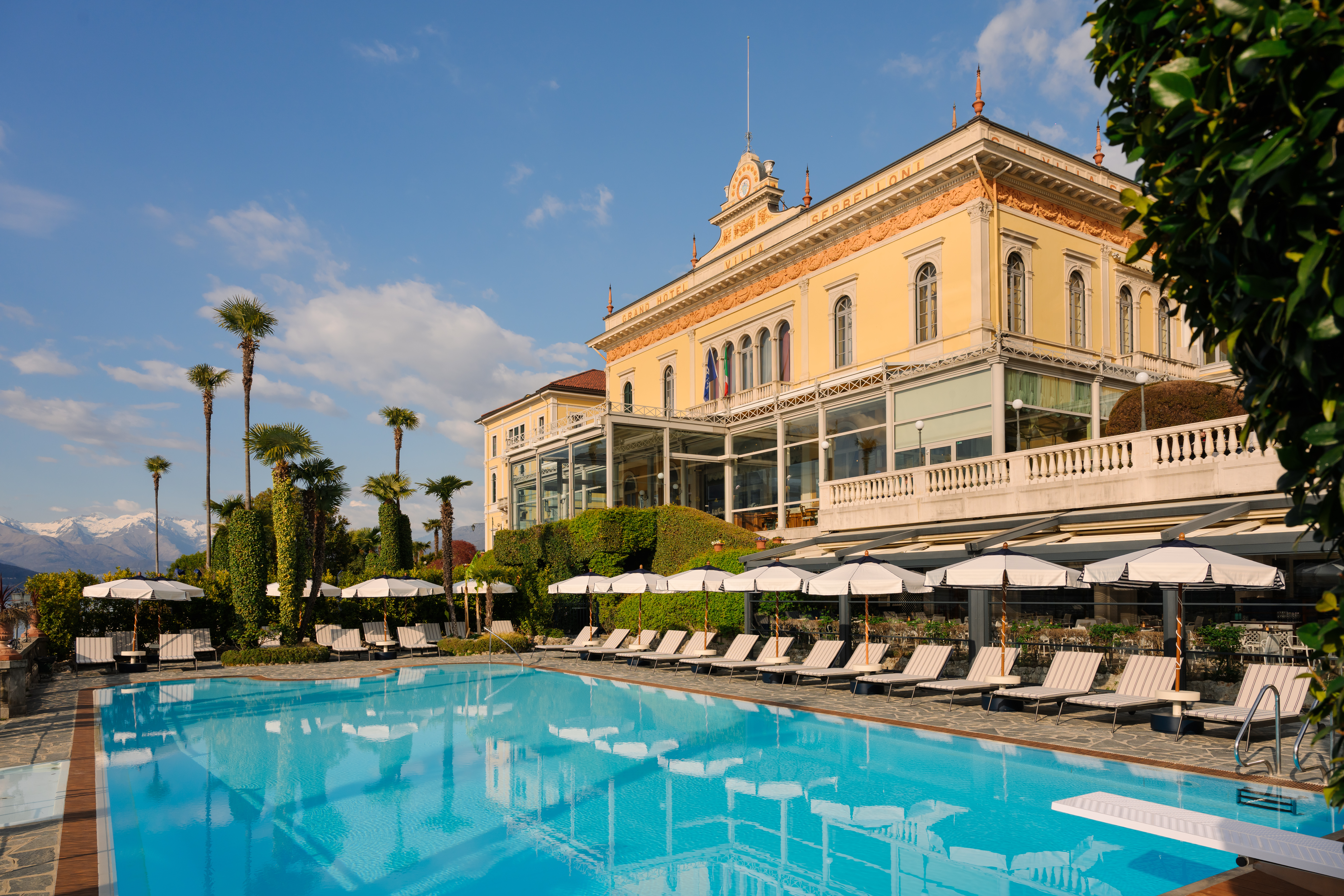 Grand Hotel Villa Serbelloni exterior, with yellow stucco walls, arched windows, restaurant covered terrace, and pool with sun loungers in the foreground