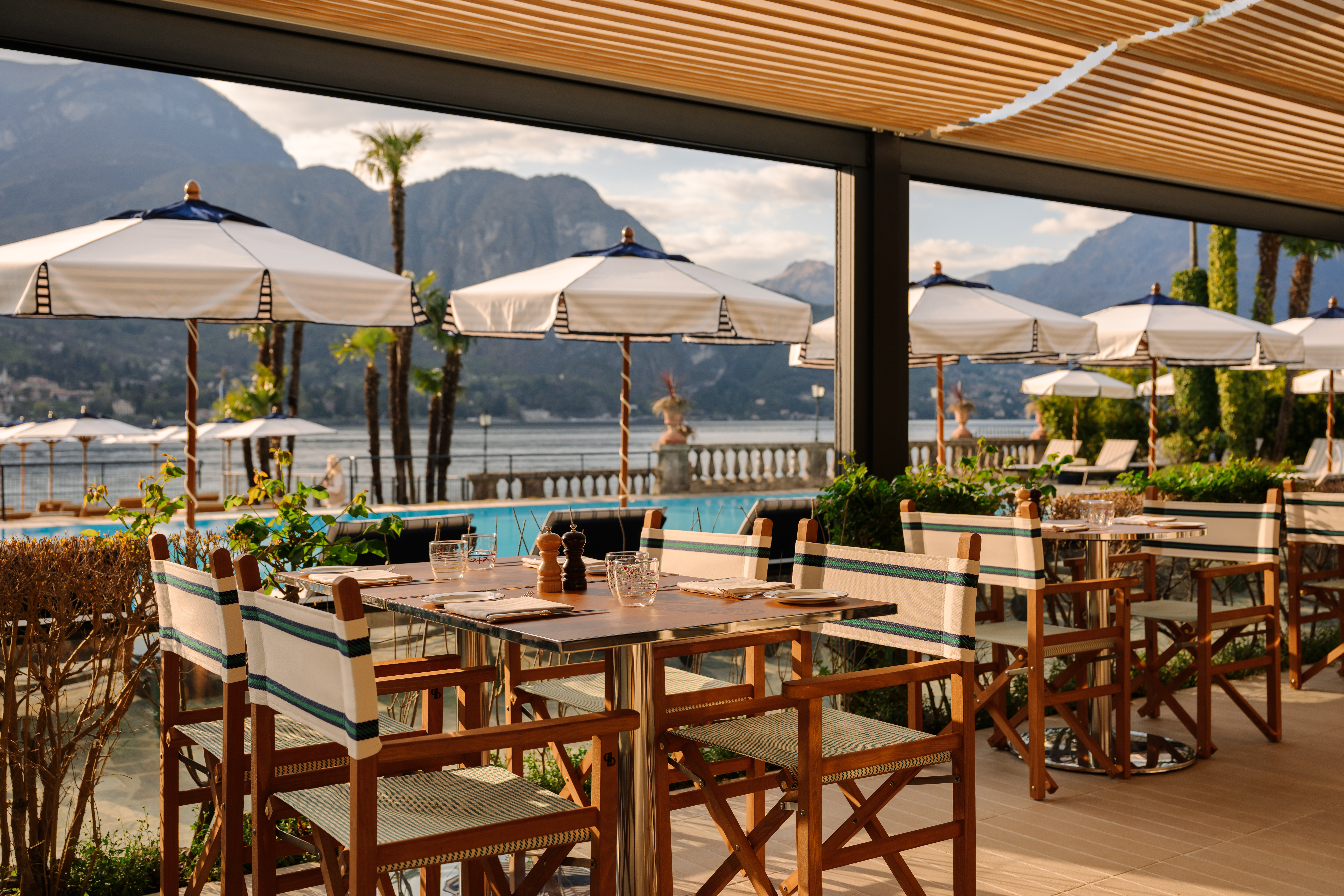 Grand Hotel Villa Serbelloni La Goletta restaurant with wooden outdoor terrace, retractable canopy, white umbrellas, and pool and lake views