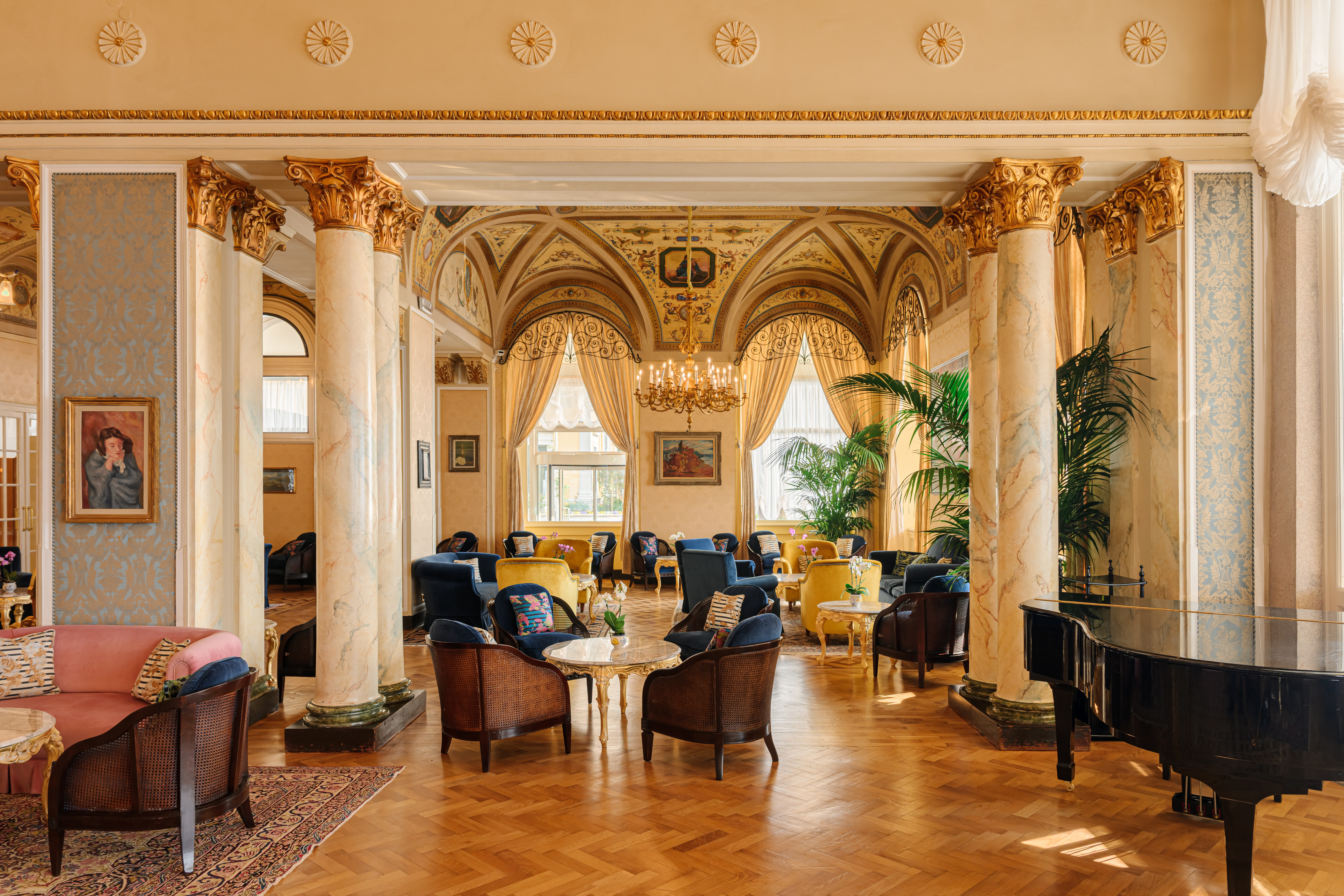 Grand Hotel Villa Serbelloni lobby with cream walls, parquet flooring, and columns topped with ornate gold decor; with a grand piano to the side