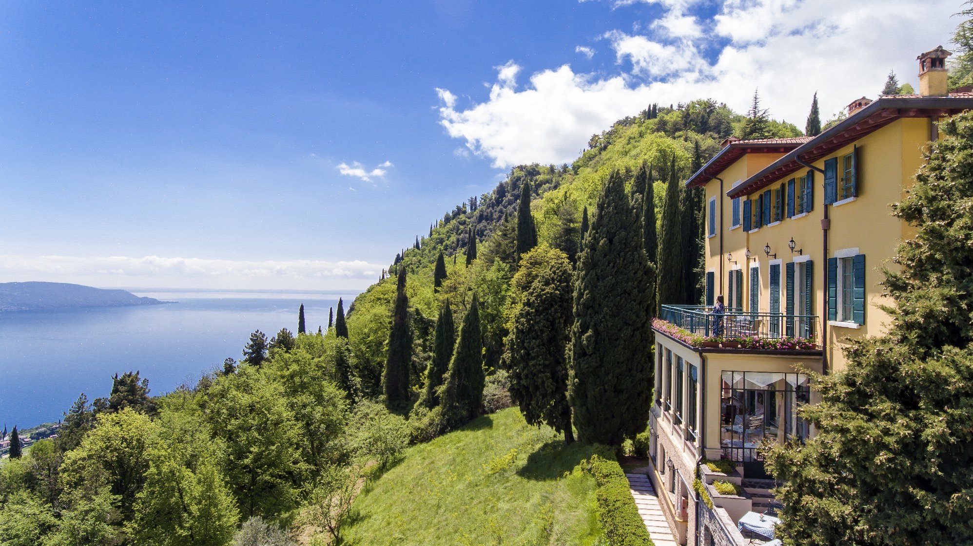 Villa Sostaga exterior with soft yellow stucco walls and green shutters, overlooking the green lakeside and the serene lake waters