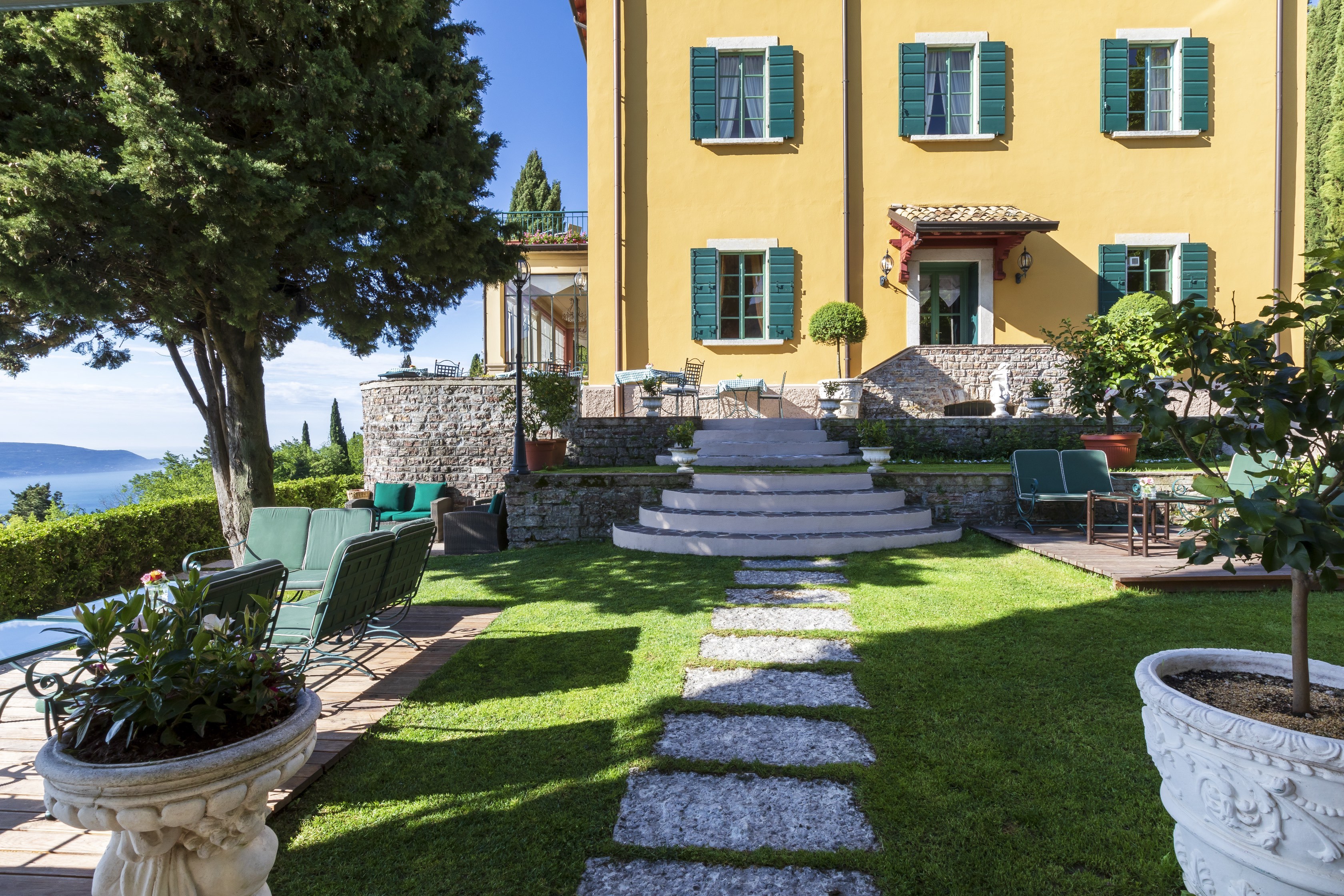 Villa Sostaga facade, with yellow-stucco walls, green shutters, lawn with paving stone pathway, and views across the lake from under the trees