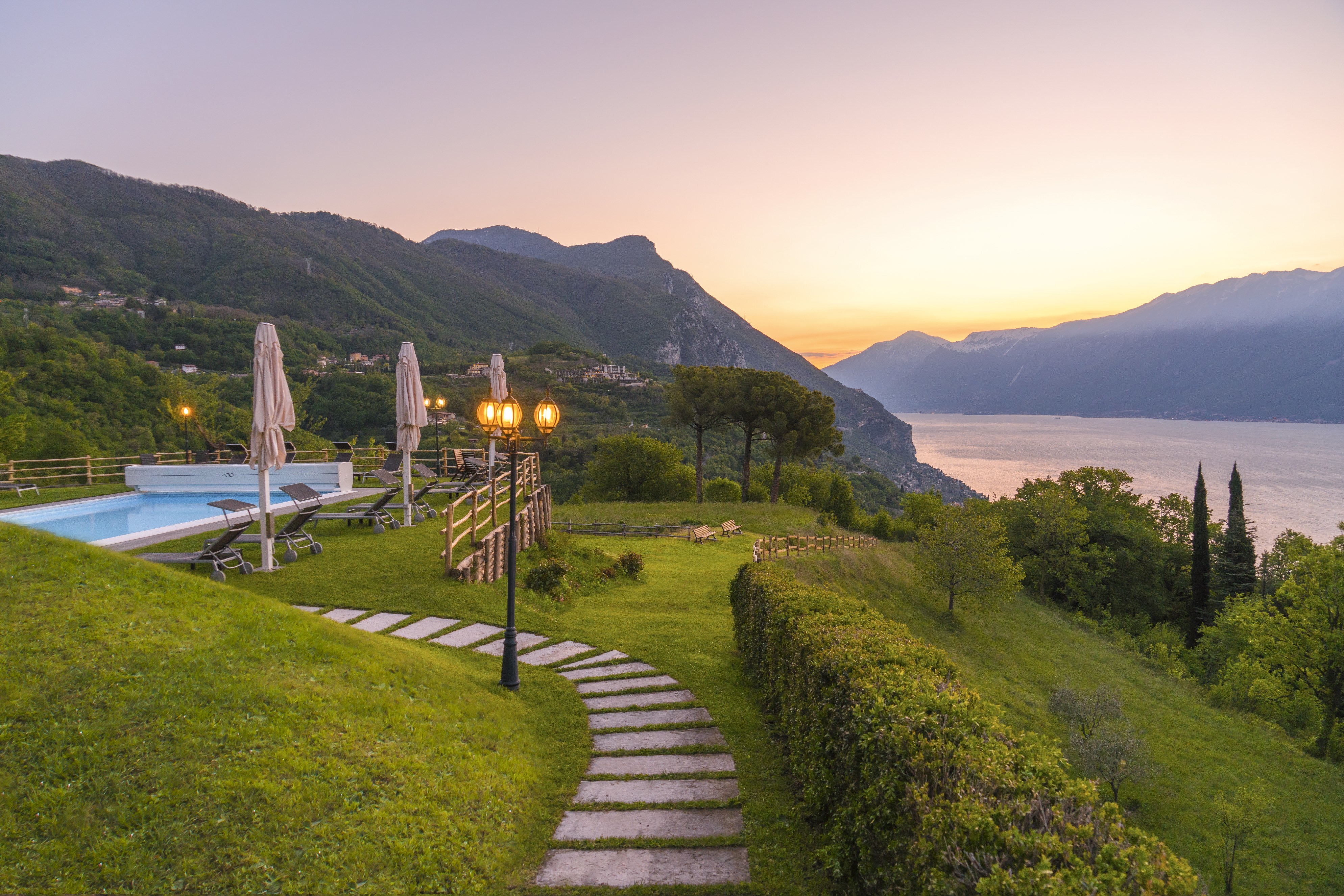 Villa Sostaga garden, showing grass and winding pathway leading to the pool, with lamps lighting the way and the view of the lake in the background