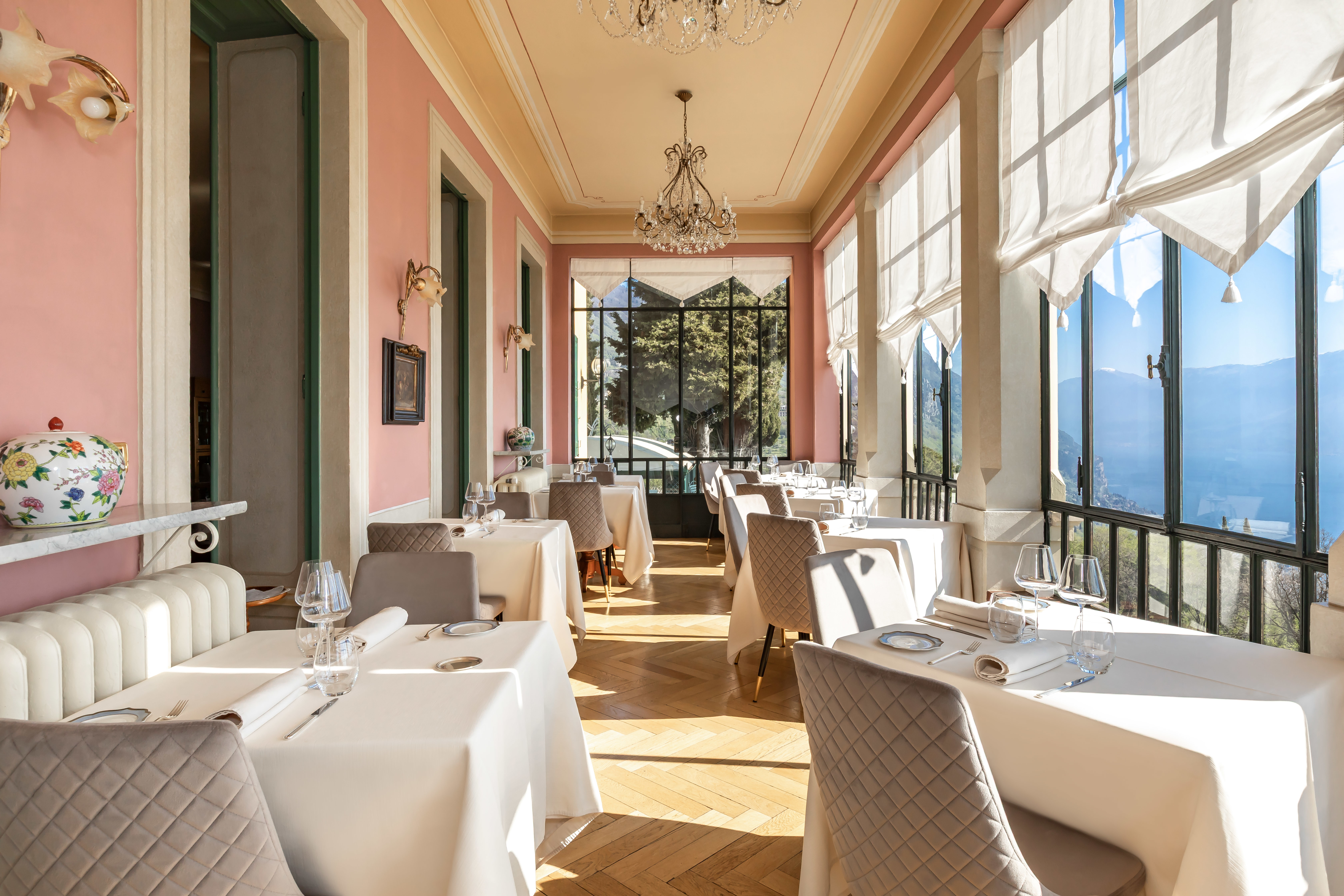 Villa Sostaga restaurant veranda, with parquet floor, white tables and chairs, pink walls, and soft linen blinds shading the views of the lake