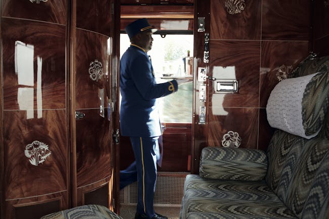 Shiny wooden train cabin with large seat and waiter with drinks on the Orient Express