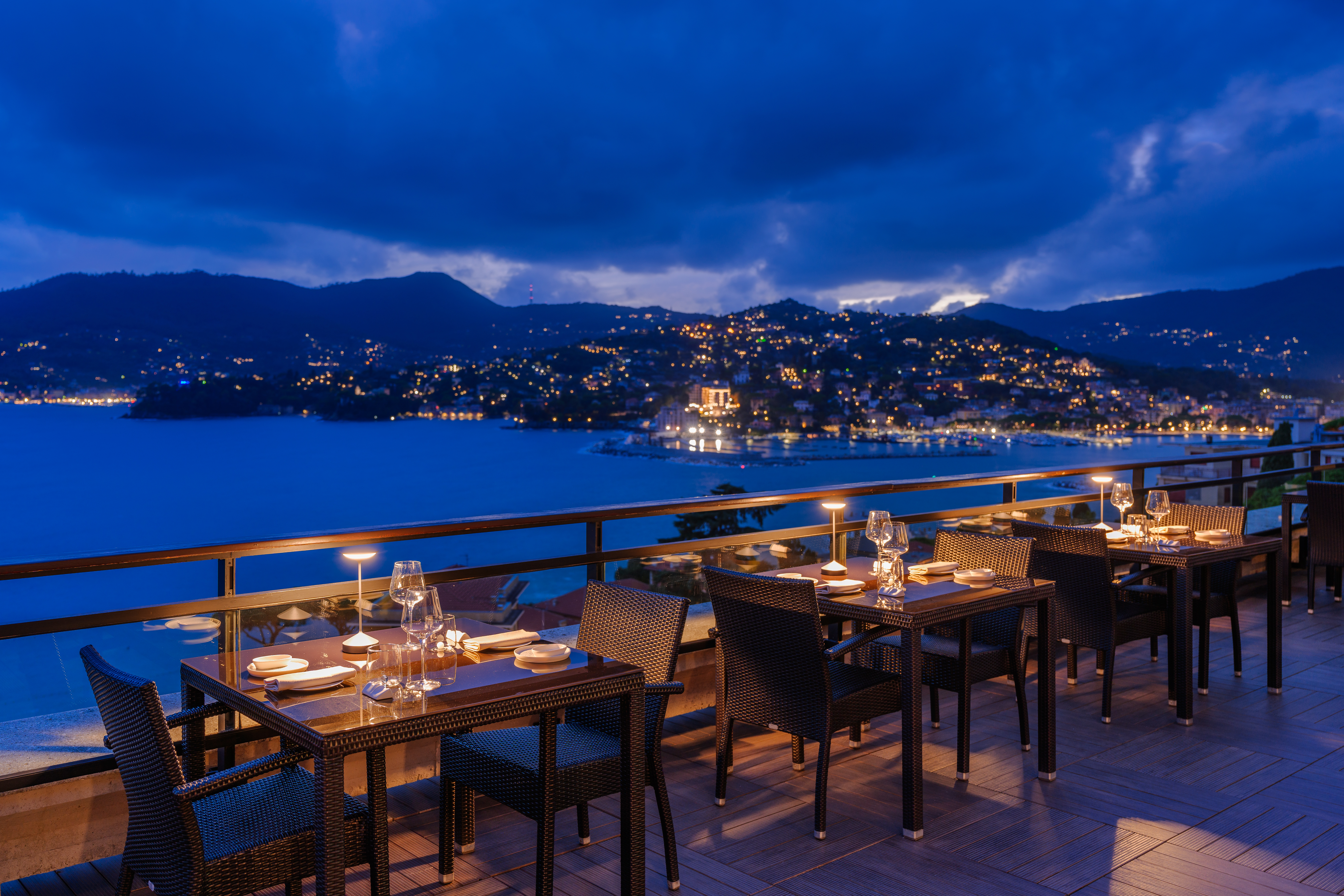 Grand Hotel Bristol Resort & Spa Le Cupole Restaurant terrace in the evening, with lamps on each table and a view out across the sea to the colourful towns and hillsides in the distance
