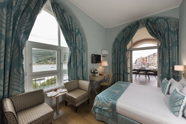 Cloister junior suite with blue colour scheme, large double bed, arm chairs and double doors opening out onto terrace balcony with views over ocean and city 