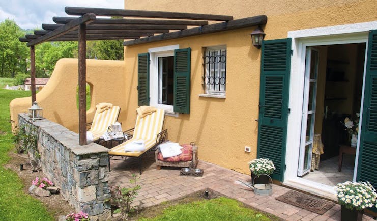 Garden patio with lounger chairs and grass outside open French window with green shutters at La Meridiana