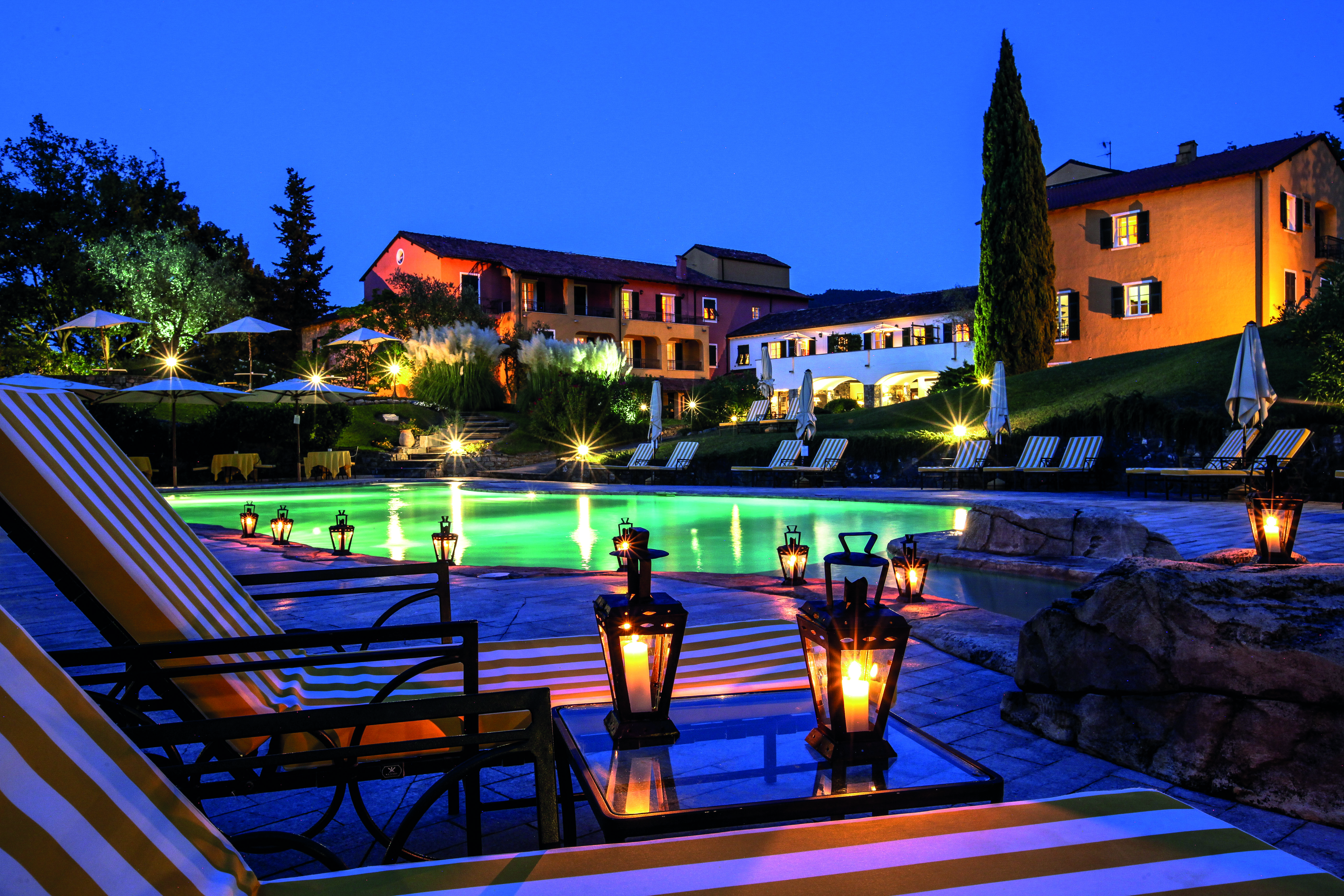 Pool with illuminated hotel buildings at night with cypress trees and lanterns on tables at La Meridiana Liguria