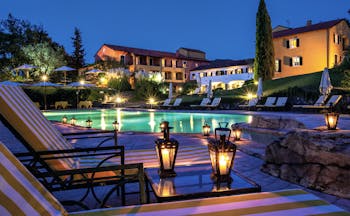 Pool with illuminated hotel buildings at night with cypress trees and lanterns on tables at La Meridiana Liguria