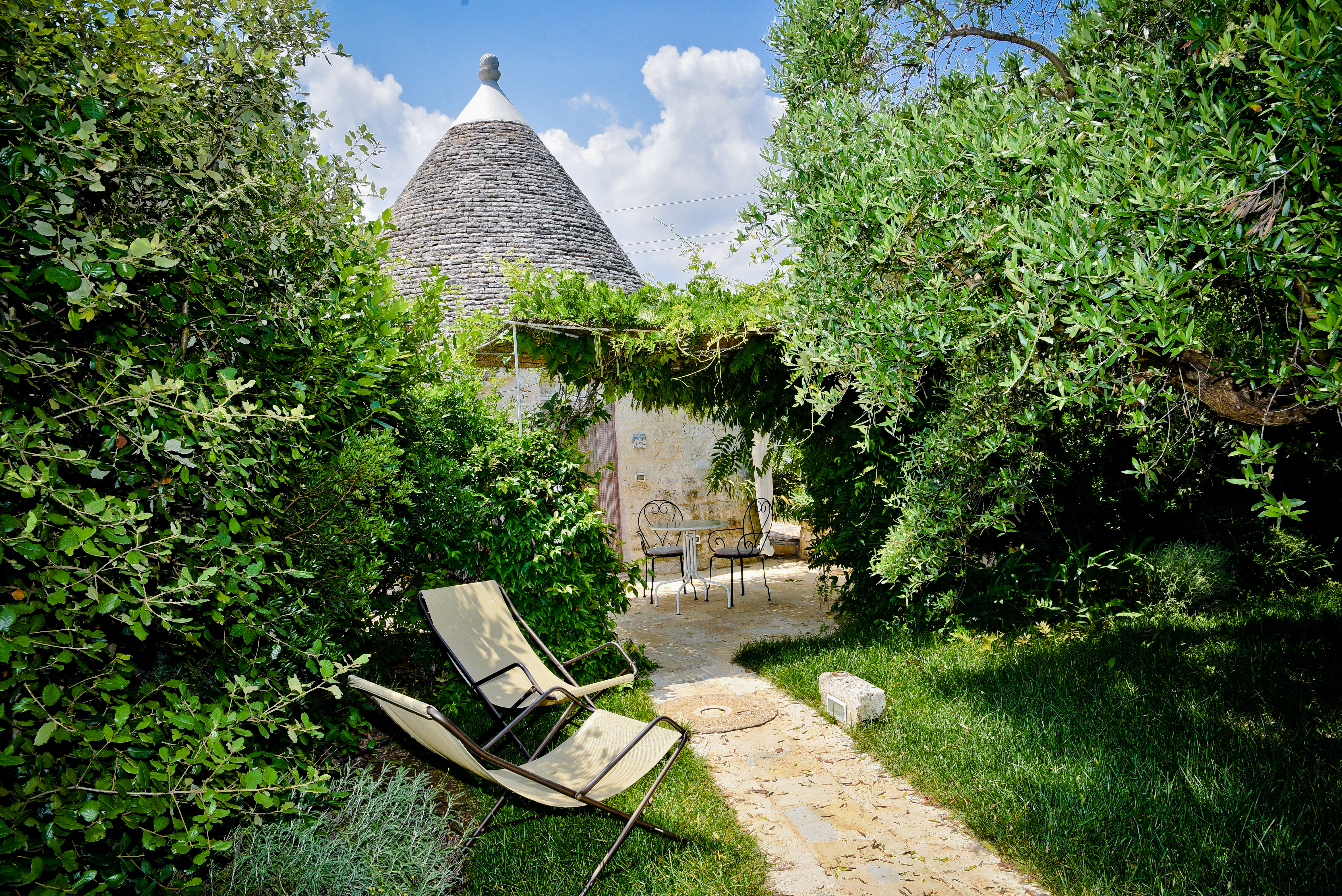 Leonardo Trulli Resort garden entrance to a trullo room, with plentiful trees and bushes, two rattan chairs, and the roof of a trullo rising behind the vine canopy