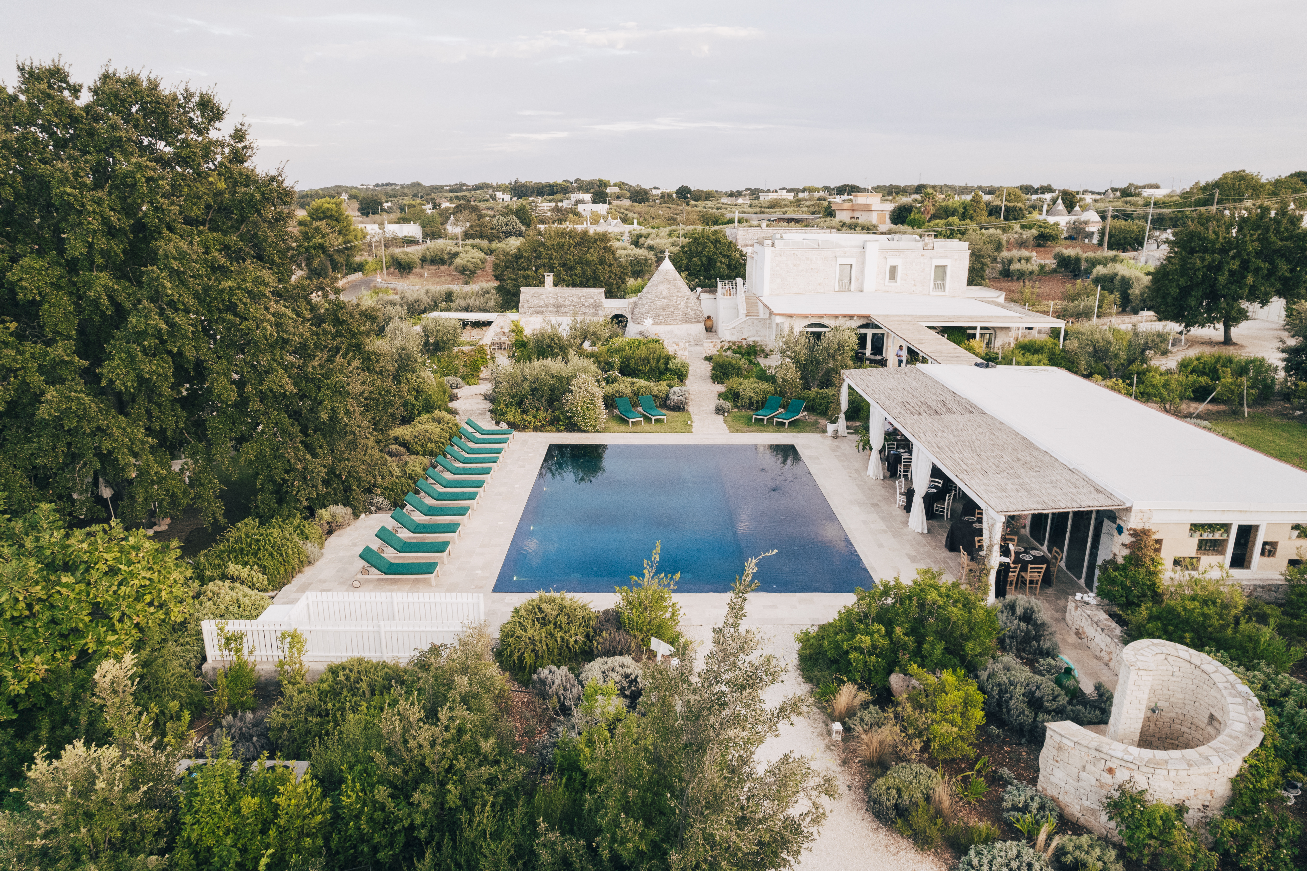 Leonardo Trulli Resort swimming pool, seen from an elevated angle, with blue loungers on a light stone patio, and a pool bar building to one side, with trees and bushes all around and trulli in the distance