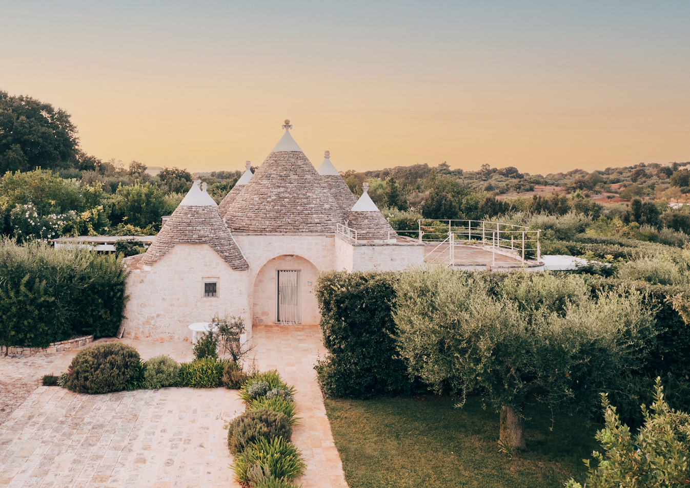 Leonardo Trulli Resort trullo room, with multiple coned roofs, pathway leading up to it, and shurbs and tree all around