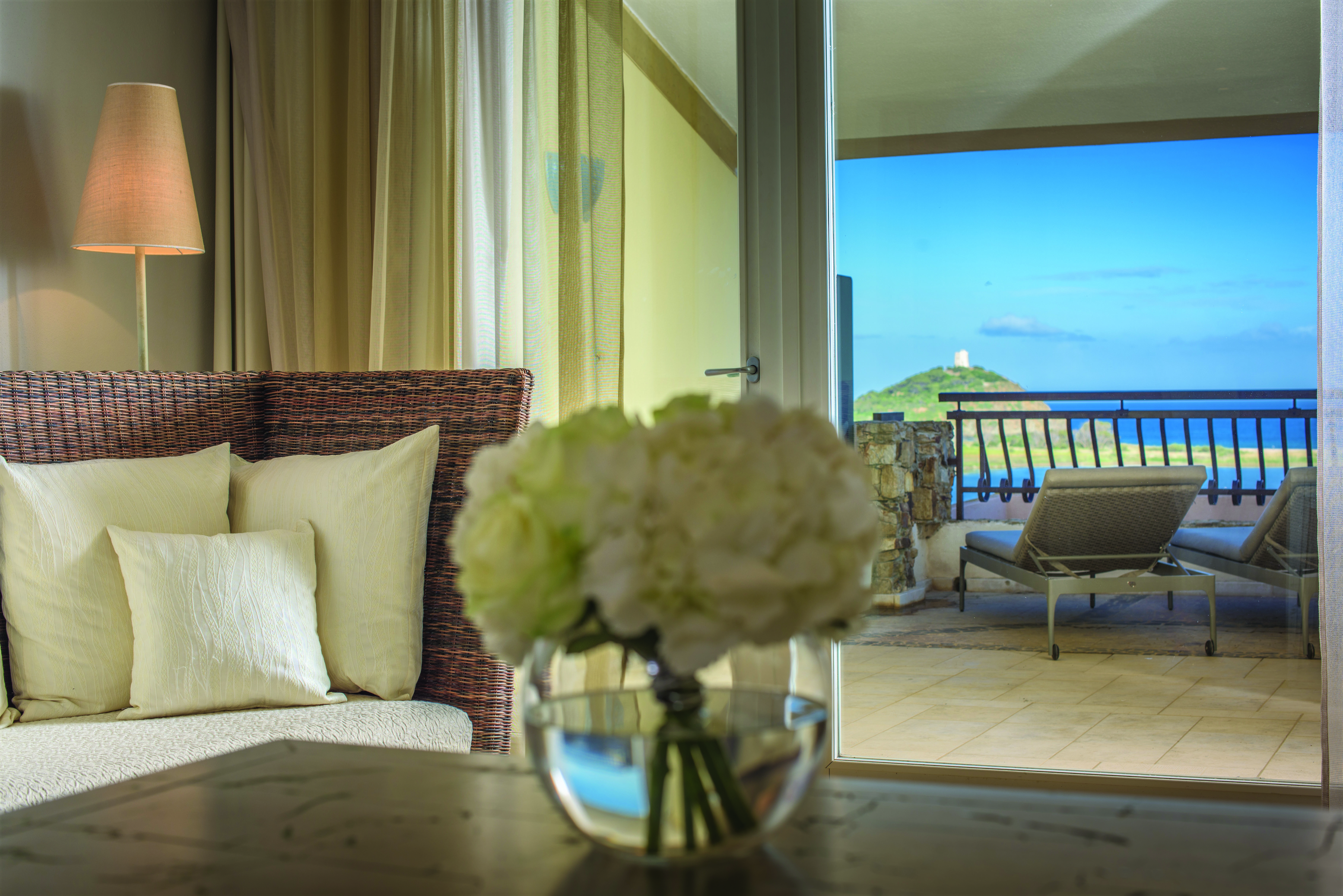 Inside the living space of a deluxe room at the Chia Laguna with white flowers on the coffee table and doors opening up onto a terrace balcony overlooking the sea 