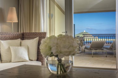 Inside the living space of a deluxe room at the Chia Laguna with white flowers on the coffee table and doors opening up onto a terrace balcony overlooking the sea 