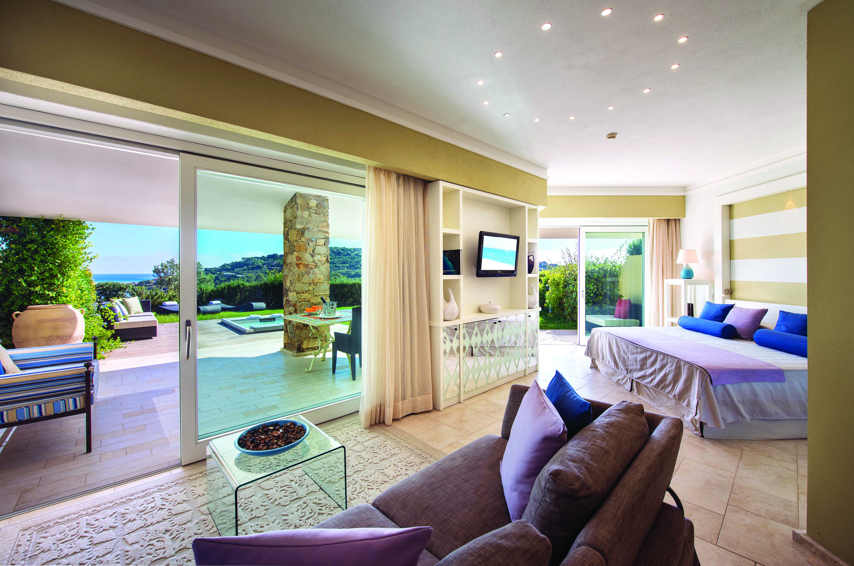 View of a Chia Laguna Garden suite with a sofa, large bed, with bi-folding doors opening onto a terrace garden