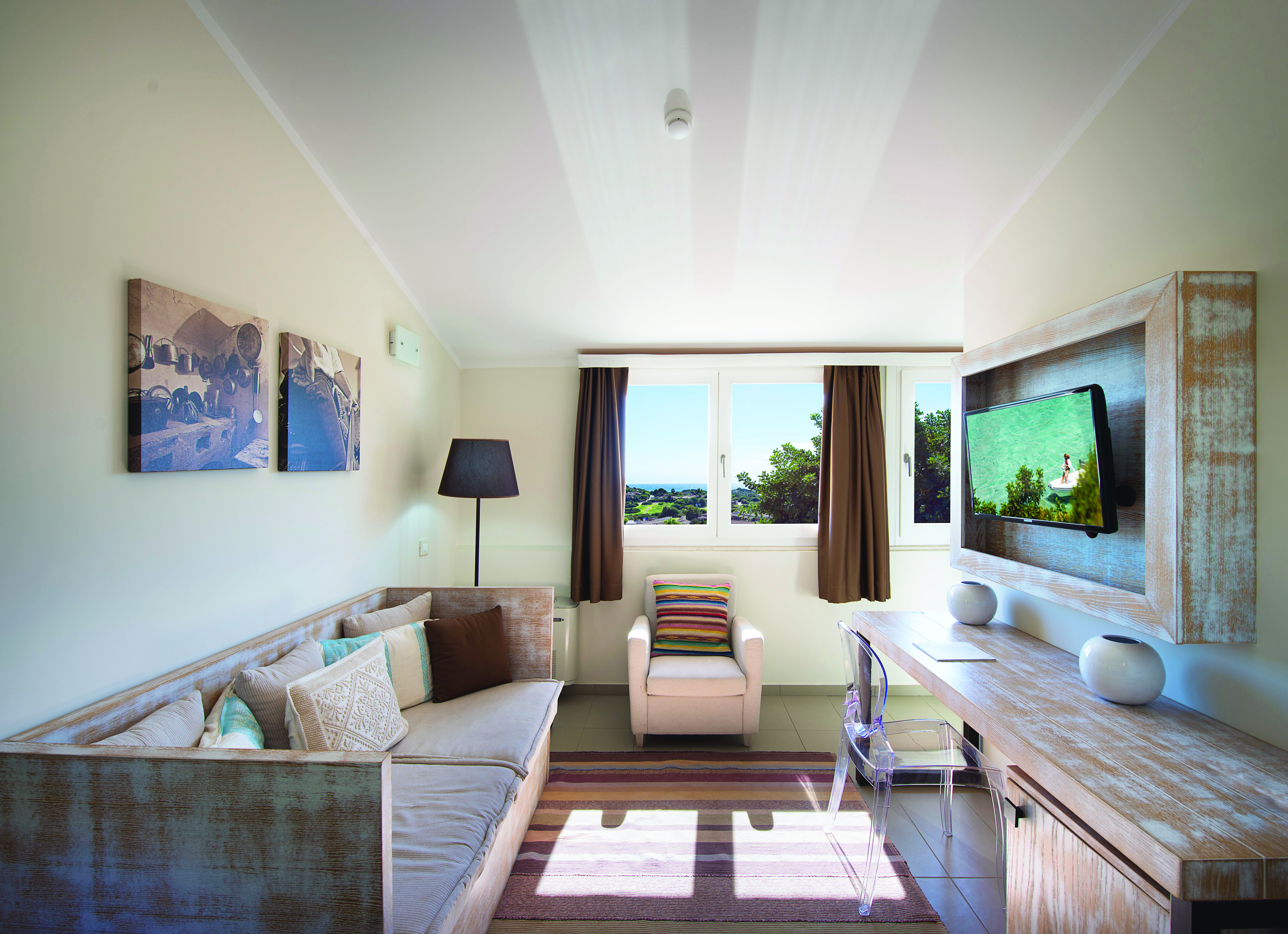 Living area with a long sofa, armcahir, wooden desk and television with a brown and cream colour scheme