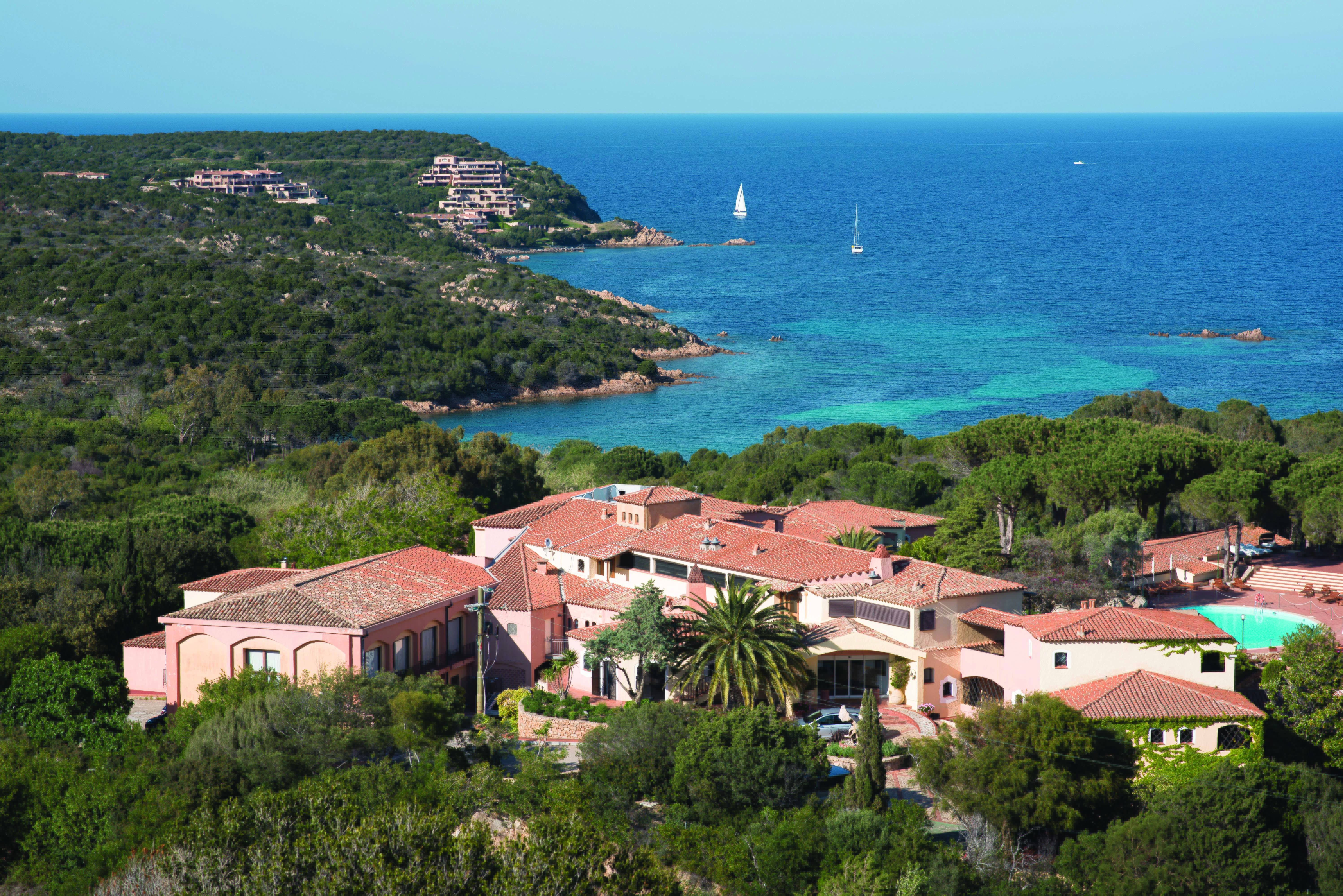 Hotel Le Ginestre Sardinia resort, hotel buildings nestled on wooded coastline, sea and beach in background