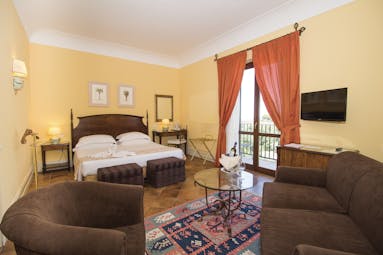 Baglio Oneto deluxe room, double bed, armchairs and sofa, tiled floor, balcony with garden views, bright traditional decor