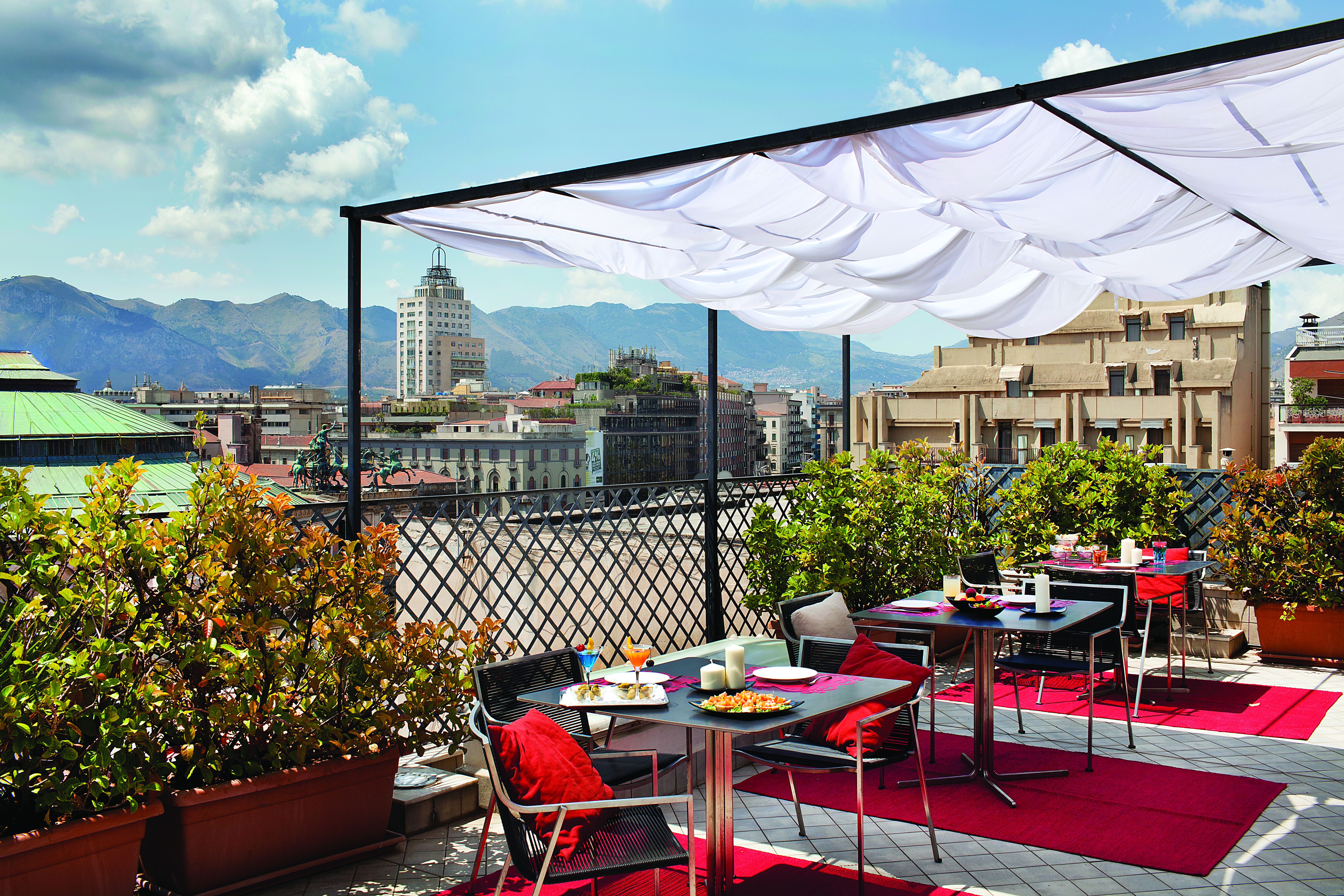 View from the rooftop terrace of Hotel Plaza Opera looking over the plaza, with red rugs and dining tables set out beneath a white veranda and amongst potted plants