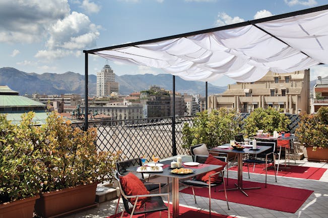View from the rooftop terrace of Hotel Plaza Opera looking over the plaza, with red rugs and dining tables set out beneath a white veranda and amongst potted plants