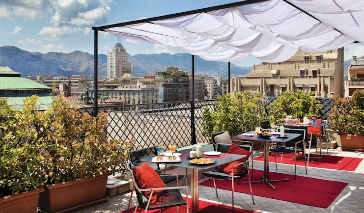 View from the rooftop terrace of Hotel Plaza Opera looking over the plaza, with red rugs and dining tables set out beneath a white veranda and amongst potted plants