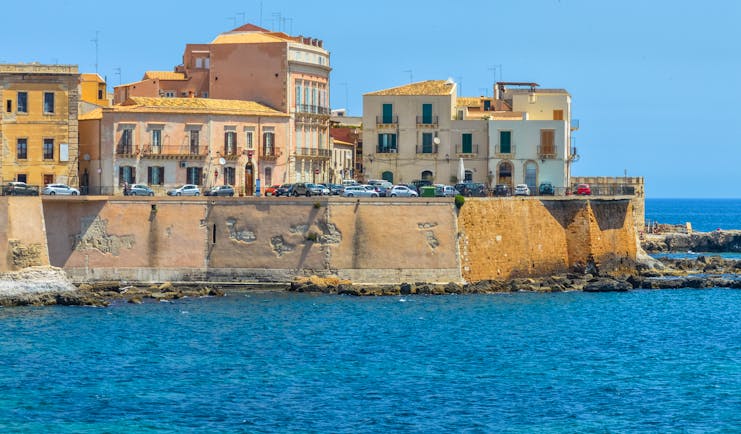 White and yellow buildings built into stone walls above the sea on the island of Ortigia in Sicily