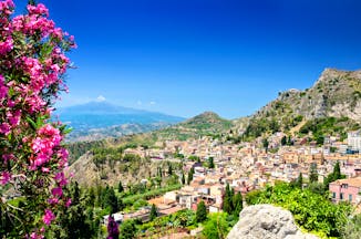 Pink flowers in foreground with town of Taormina and Mount Etna volcanic dome in background in Sicily