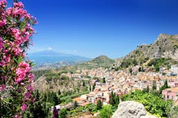 Pink flowers in foreground with town of Taormina and Mount Etna volcanic dome in background in Sicily