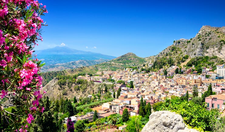 Pink flowers in foreground with town of Taormina and Mount Etna volcanic dome in background in Sicily