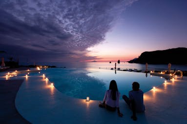 Pool at night with candles lit up around the edge of the pool and the sun setting over the sea in the distance