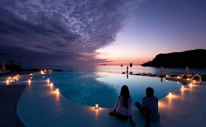 Pool at night with candles lit up around the edge of the pool and the sun setting over the sea in the distance