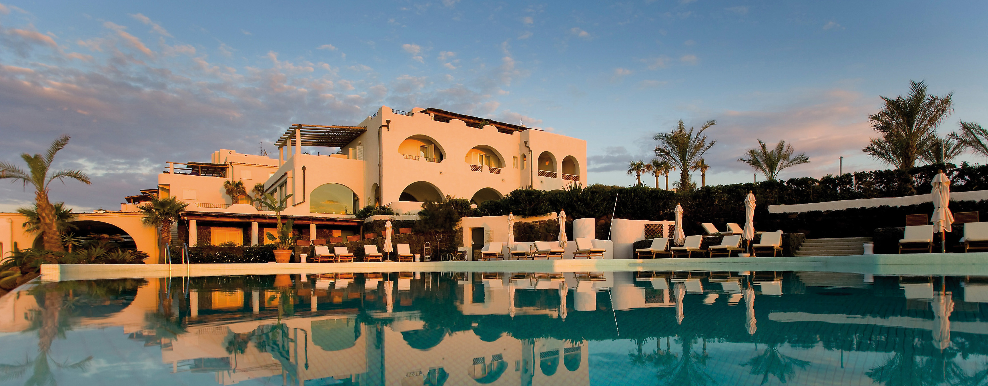 View of hotel exterior and pool infront with palm trees around and sun loungers 
