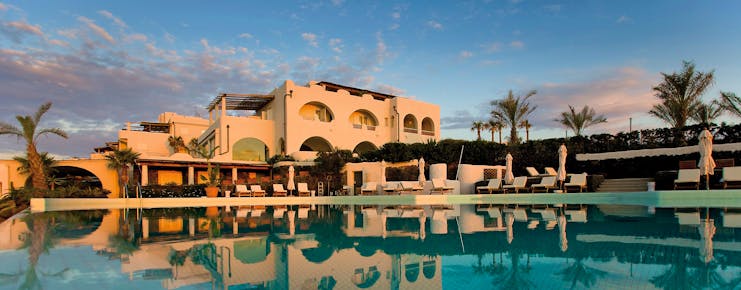 View of hotel exterior and pool infront with palm trees around and sun loungers 