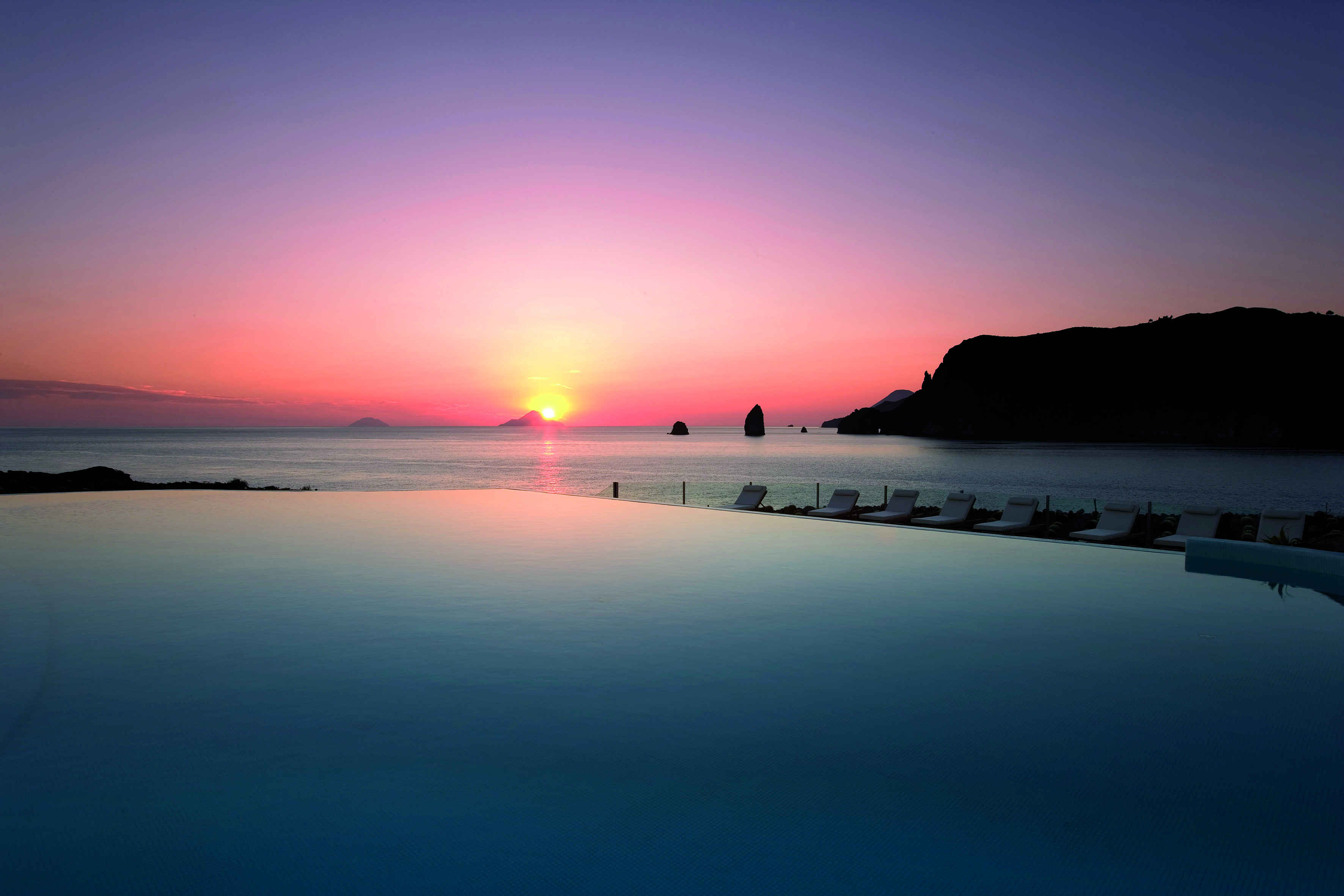 View of a pink sunset setting over the sea with the Therasia resort swimming pool in front 