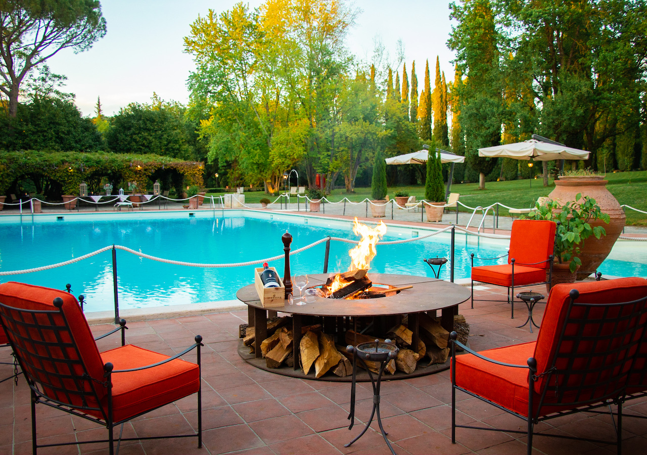 Borgo San Luigi poolside, with red cushioned wrought iron chairs set around an outdoor fire, with gardens and cypress trees in the background