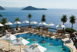 Pool view from above with sunloungers and white umbrellas set up around the pool with palm trees and a view of the ocean