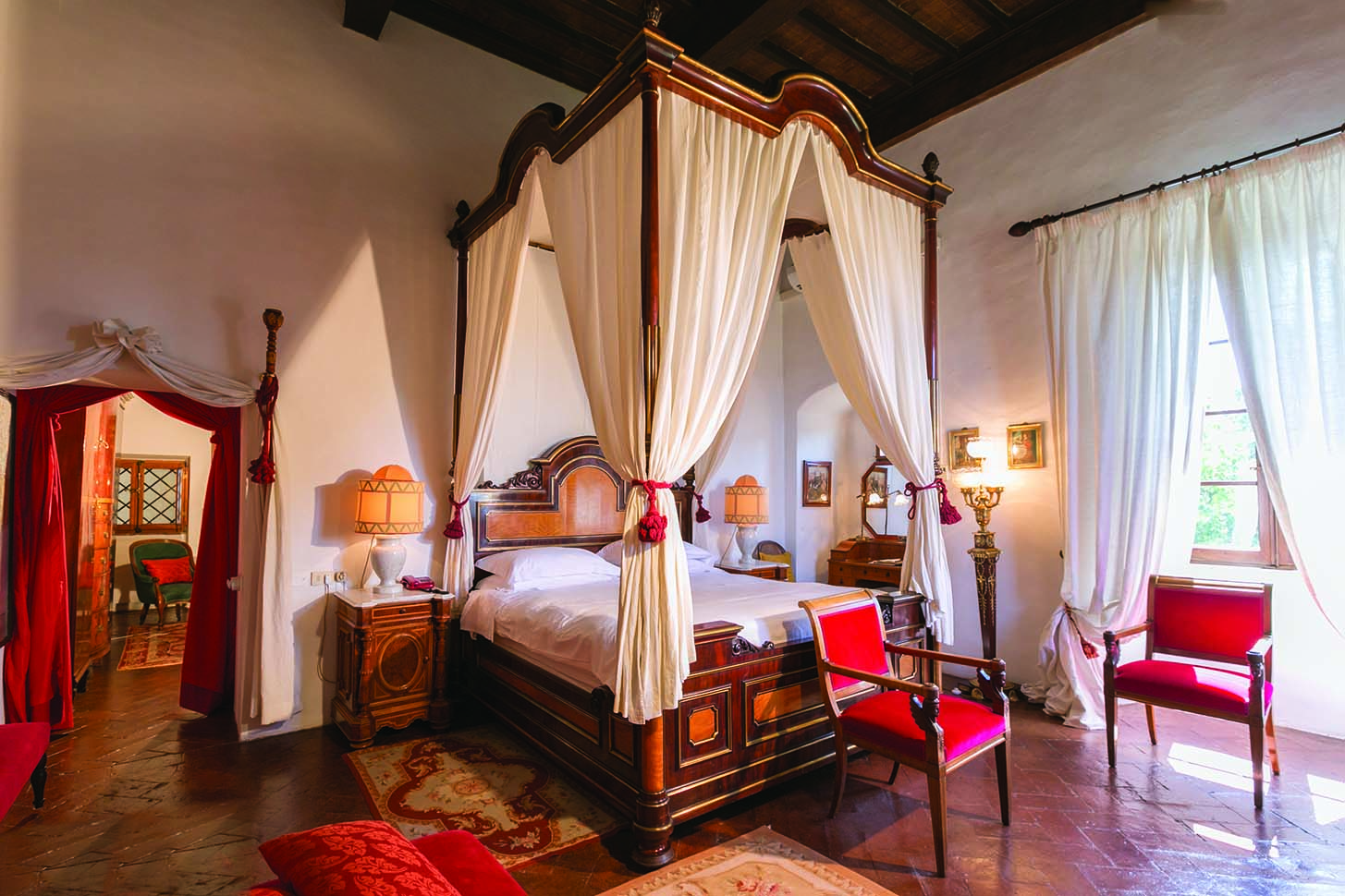 Fox suite at the Relais la Suvera with a large wooden bed with white drapes, a white and red colour scheme with red chairs around the room 