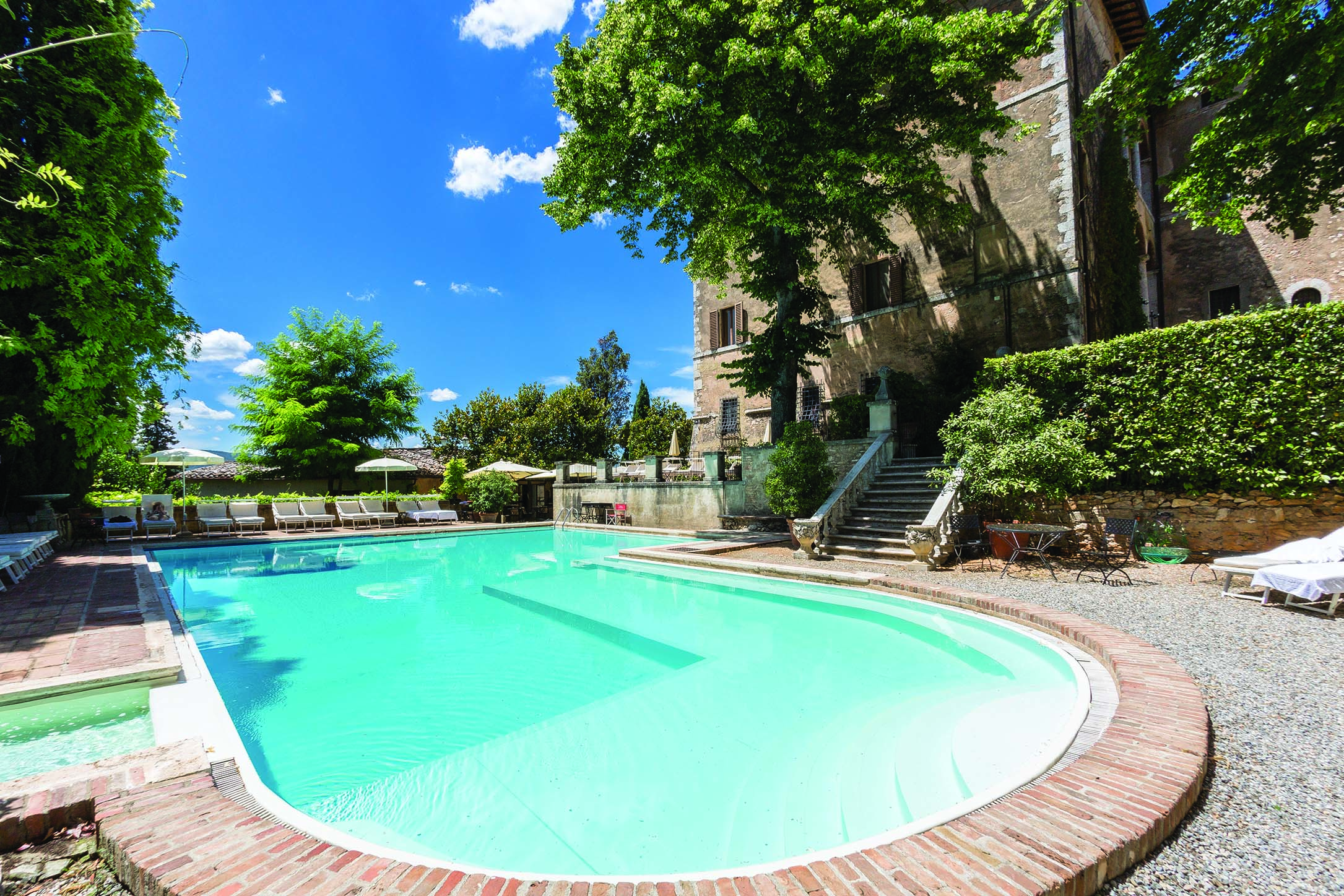View of the pool at the Relais la Suvera with greenery and white sunbeds surrounding the pool and stone stairs leading up to the hotel