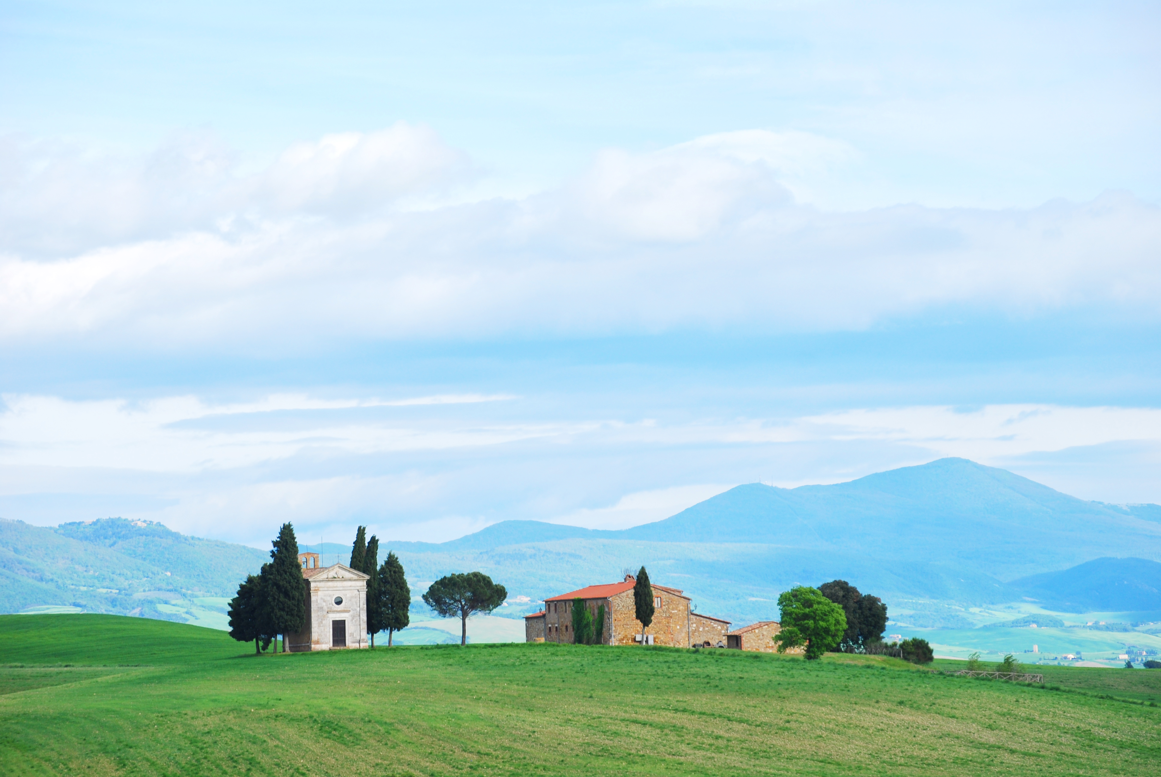 Tuscany field with small white chapel flanked by cypress trees with mountains behind