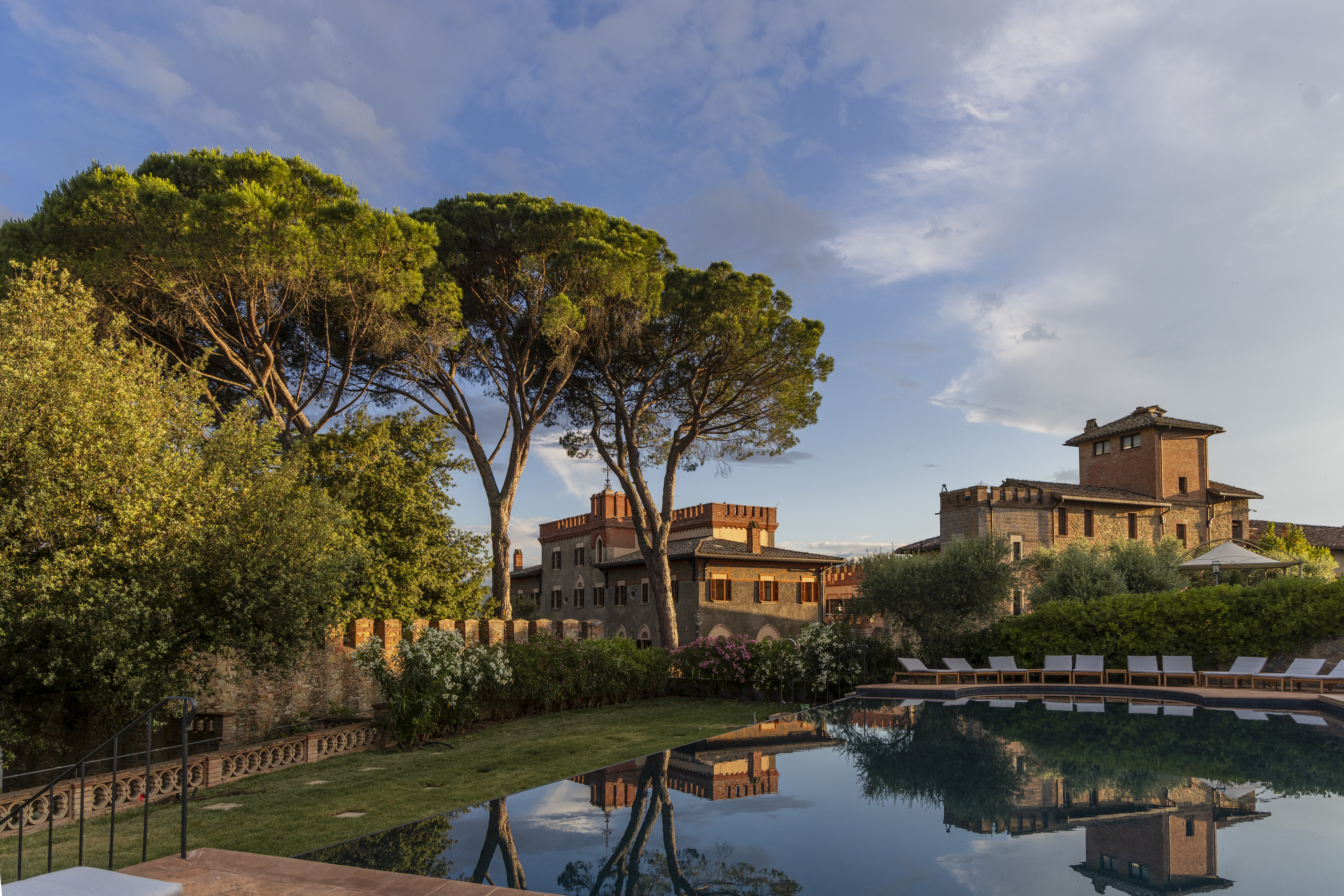 Borgo dei Conti poolside at sunset, with turquoise water lined with sun loungers, tall trees and bushes, and the ochre hotel buildings in the distance