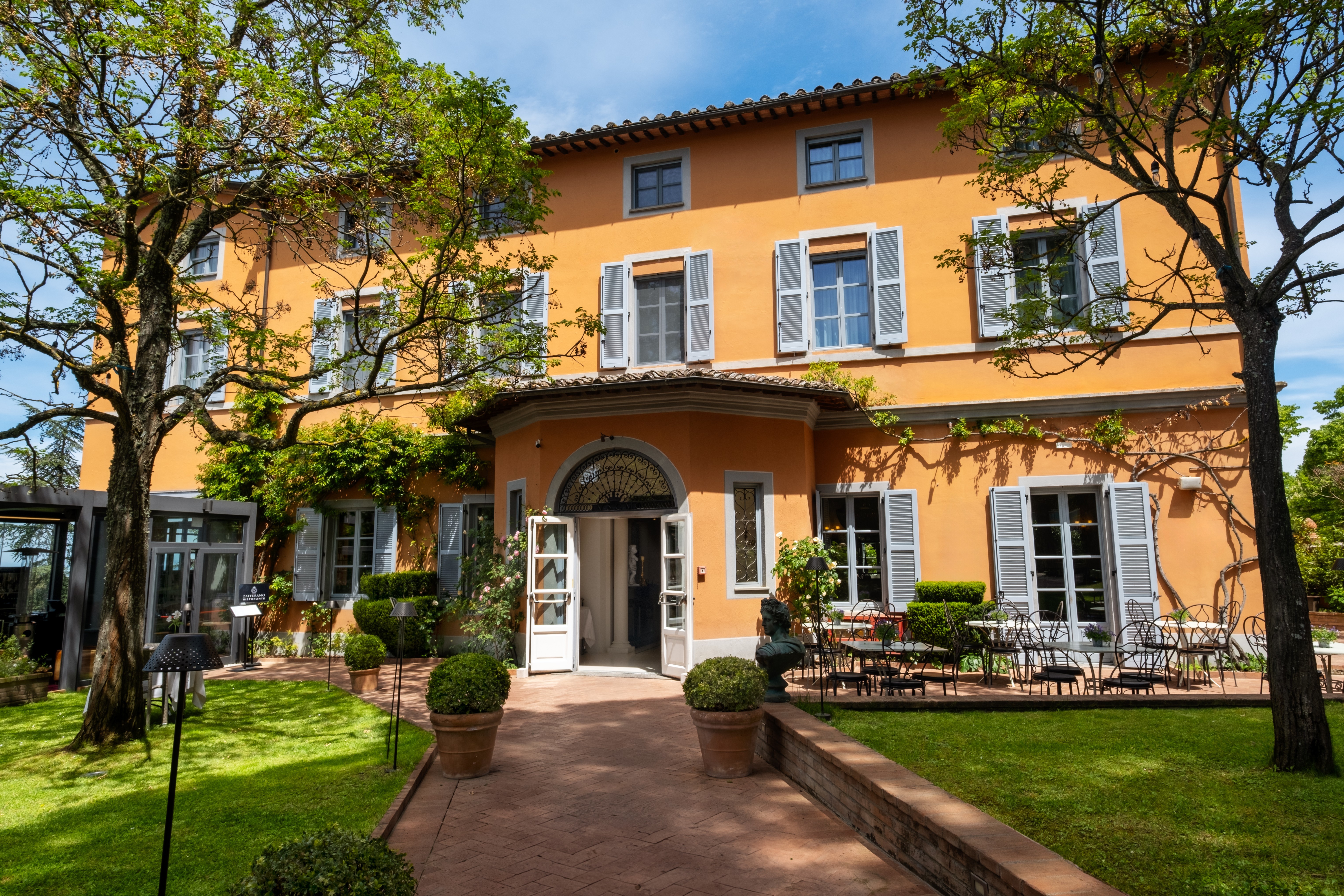 Hotel Vannucci exterior, showing yellow walls with white shuttered windows, pathway leading to the symmetrical hotel buildings, with chairs and tables on the patio and lawn