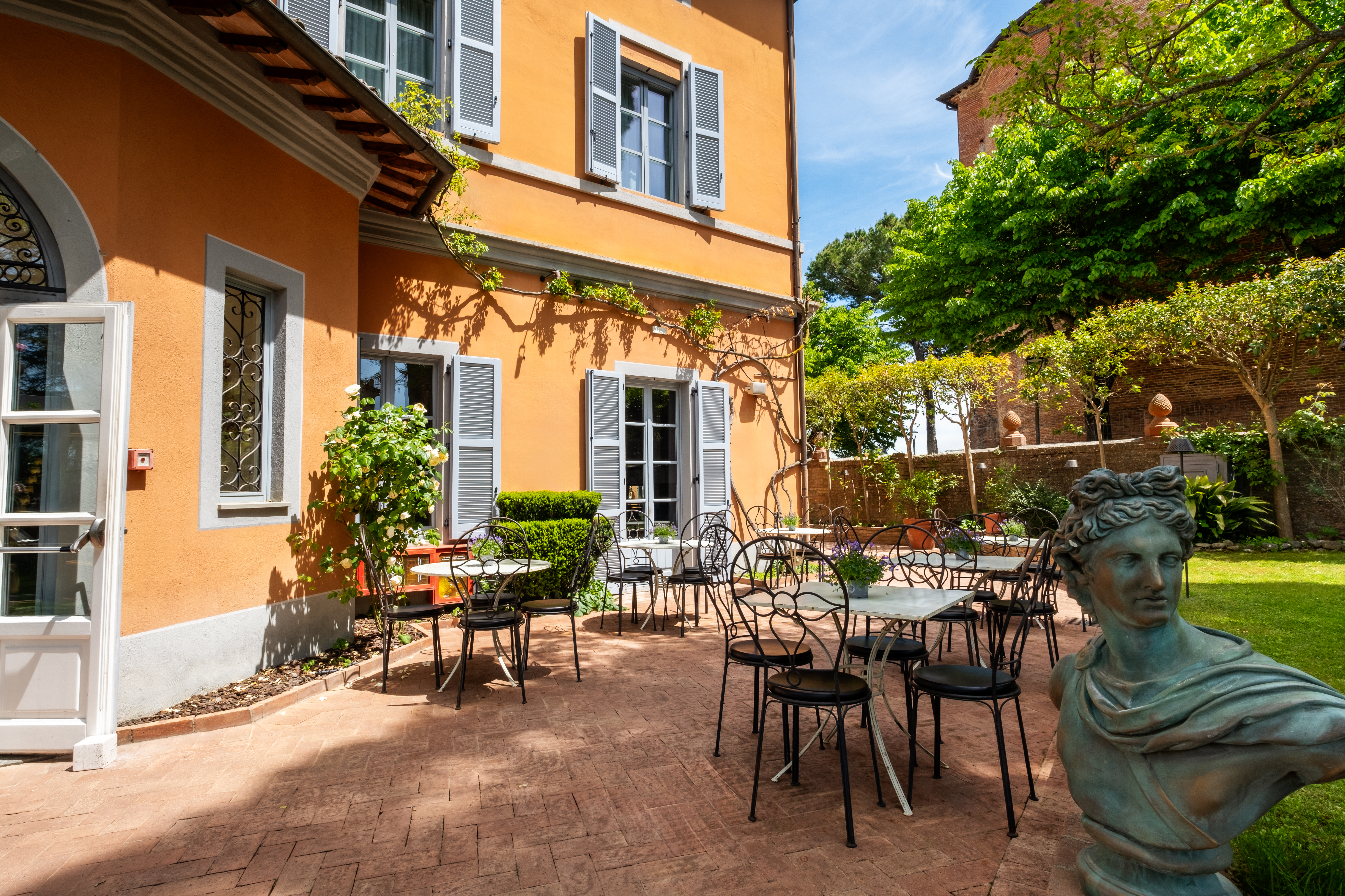 Hotel Vannucci patio with wrought iron furniture, small statues, grassy areas, and the hotel's yellow walls to the left