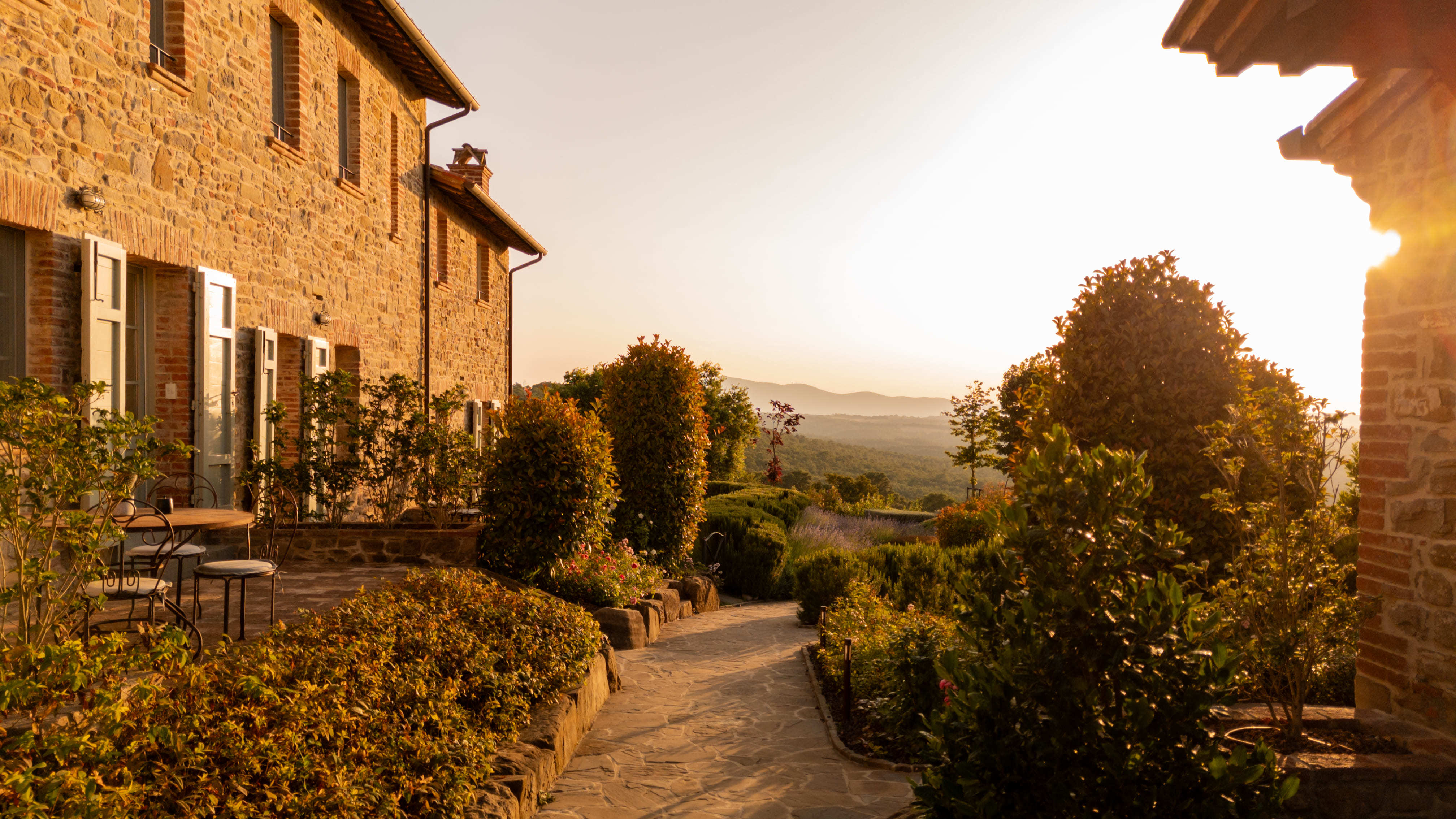 I Borghi Dell'Eremo pathway leading between hotel buildings, with wrought iron chairs and tables on one side, against the ochre stone walls and open shutters, with trees and shrubs glowing in the afternoon light