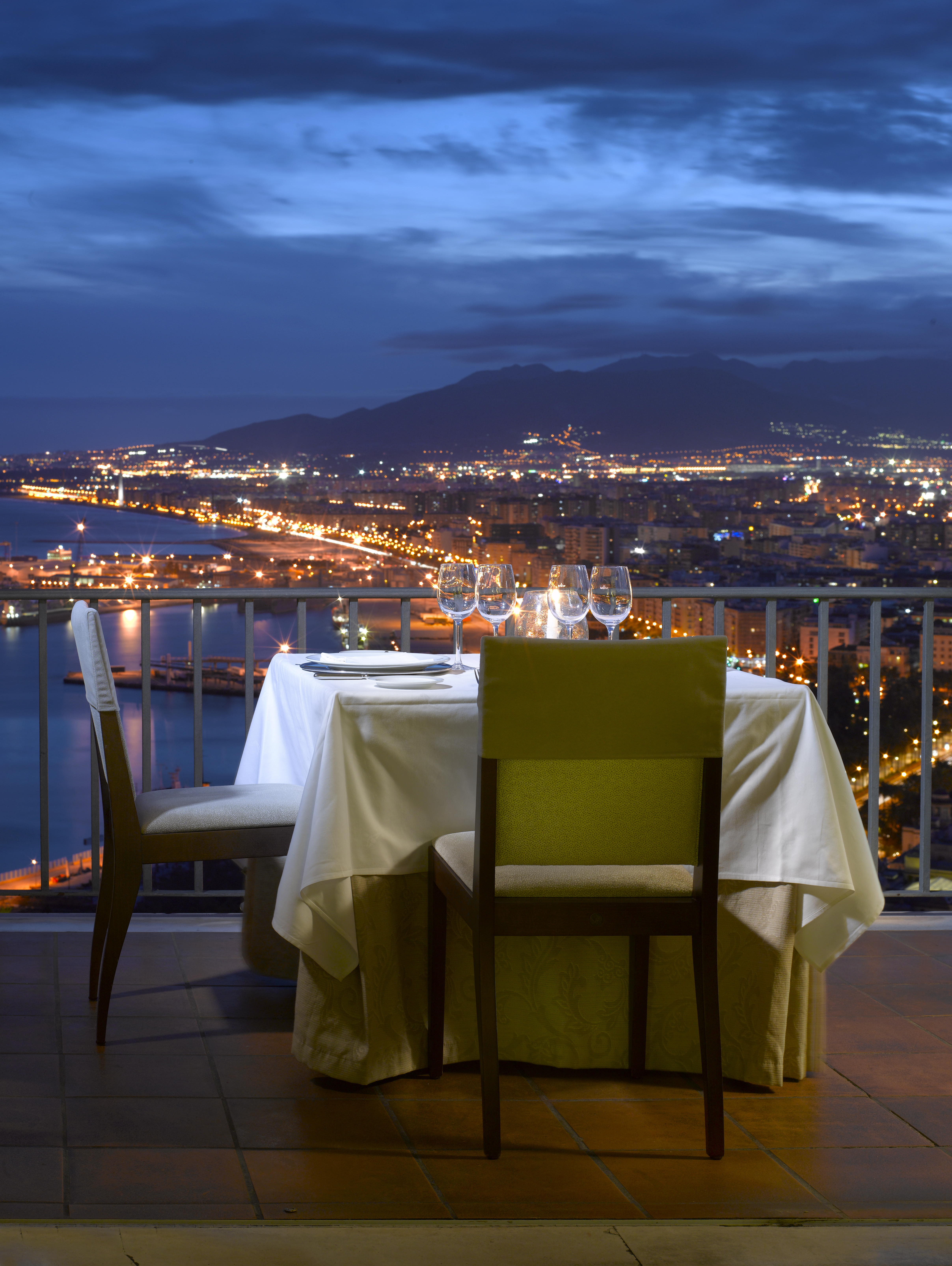 Parador de Malaga Gibralfaro dining terrace, table set for two, balcony overlooking the coast, town and mountain at night