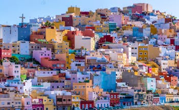 Brightly coloured houses of reds, pinks, blues and yellow on hillside with cross in sky in San Juan Las Palmas on Gran Canaria