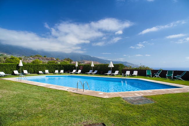 Outdoor swiming pool with grass surface nearby and white sunloungers and umbrellas set up around the edge of the pool