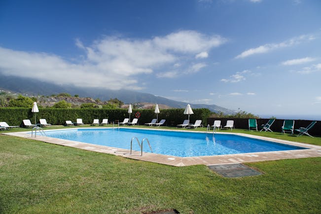 Outdoor swiming pool with grass surface nearby and white sunloungers and umbrellas set up around the edge of the pool
