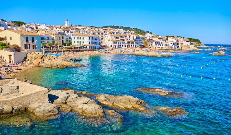 Blue sea lapping rounded rocks on coast with village behind at Palafrugell in Catalonia
