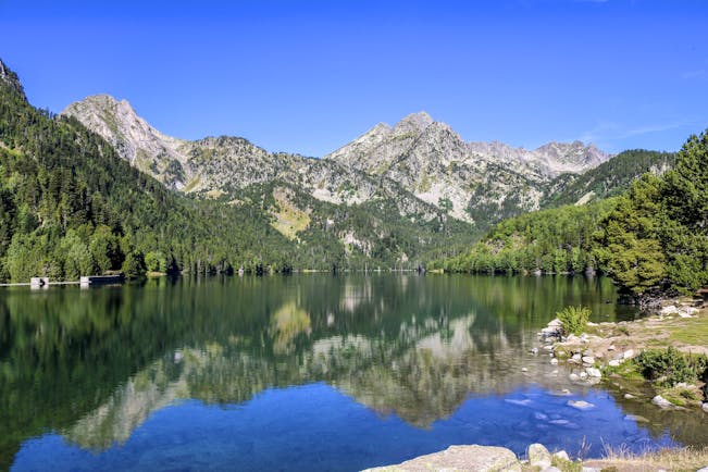 Lake with clear water surrounded by pine-clad hills and grey rocky mountains in Catalonia